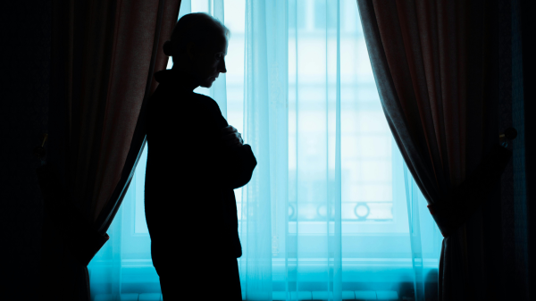 Silhouette of an older woman standing by a window, holding her arms close while thinking about a midlife shift