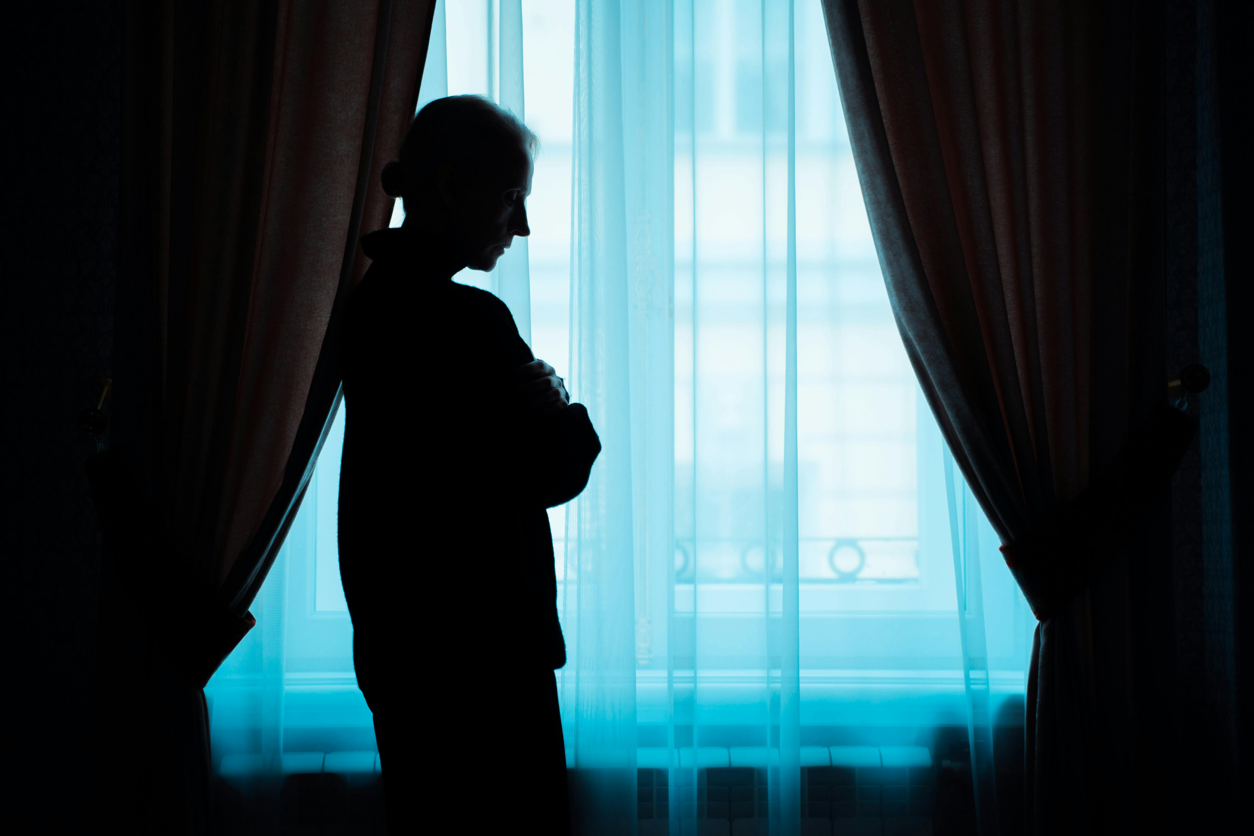 Silhouette of an older woman standing by a window, holding her arms close while thinking about a midlife shift