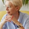 Thoughtful woman in her 40s with short blonde hair and glasses, resting her chin on her hands in a calm, modern setting