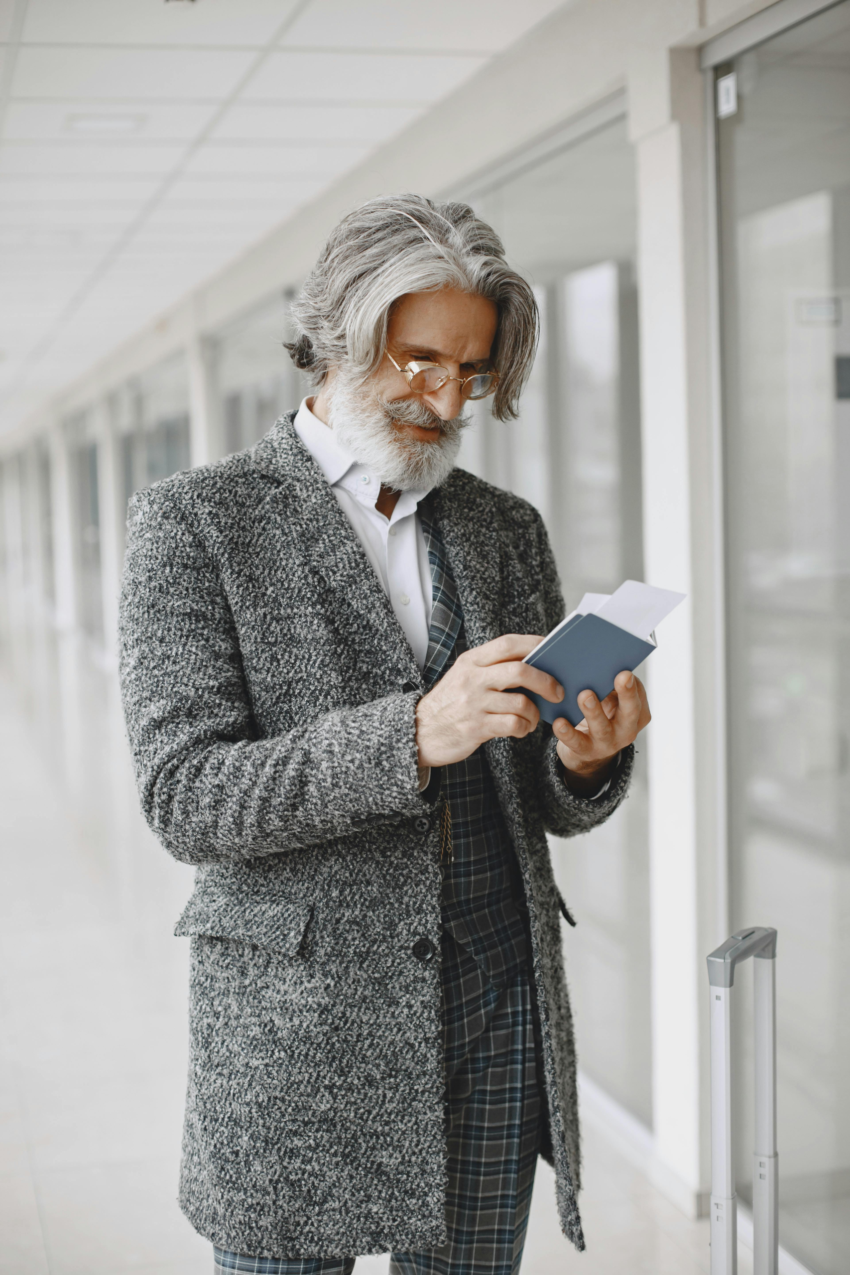 Older man reading a book while standing with luggage in a modern airport hallway