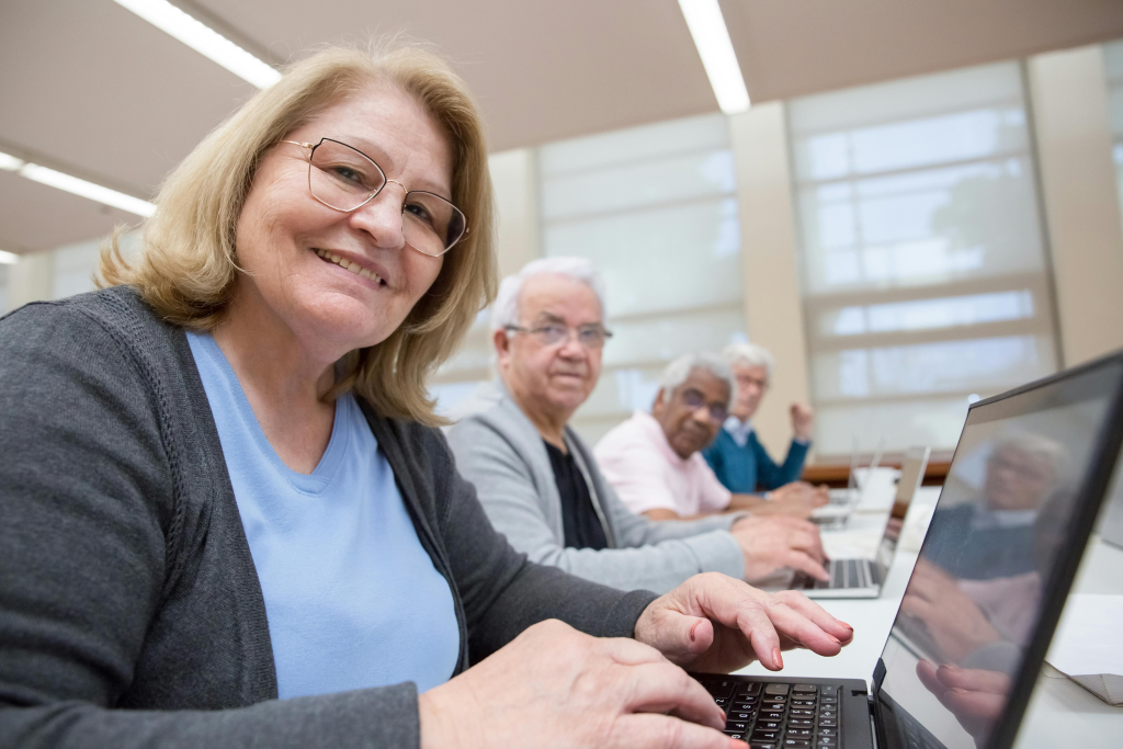 Older woman smiling while using a laptop in a classroom with other seniors learning technology