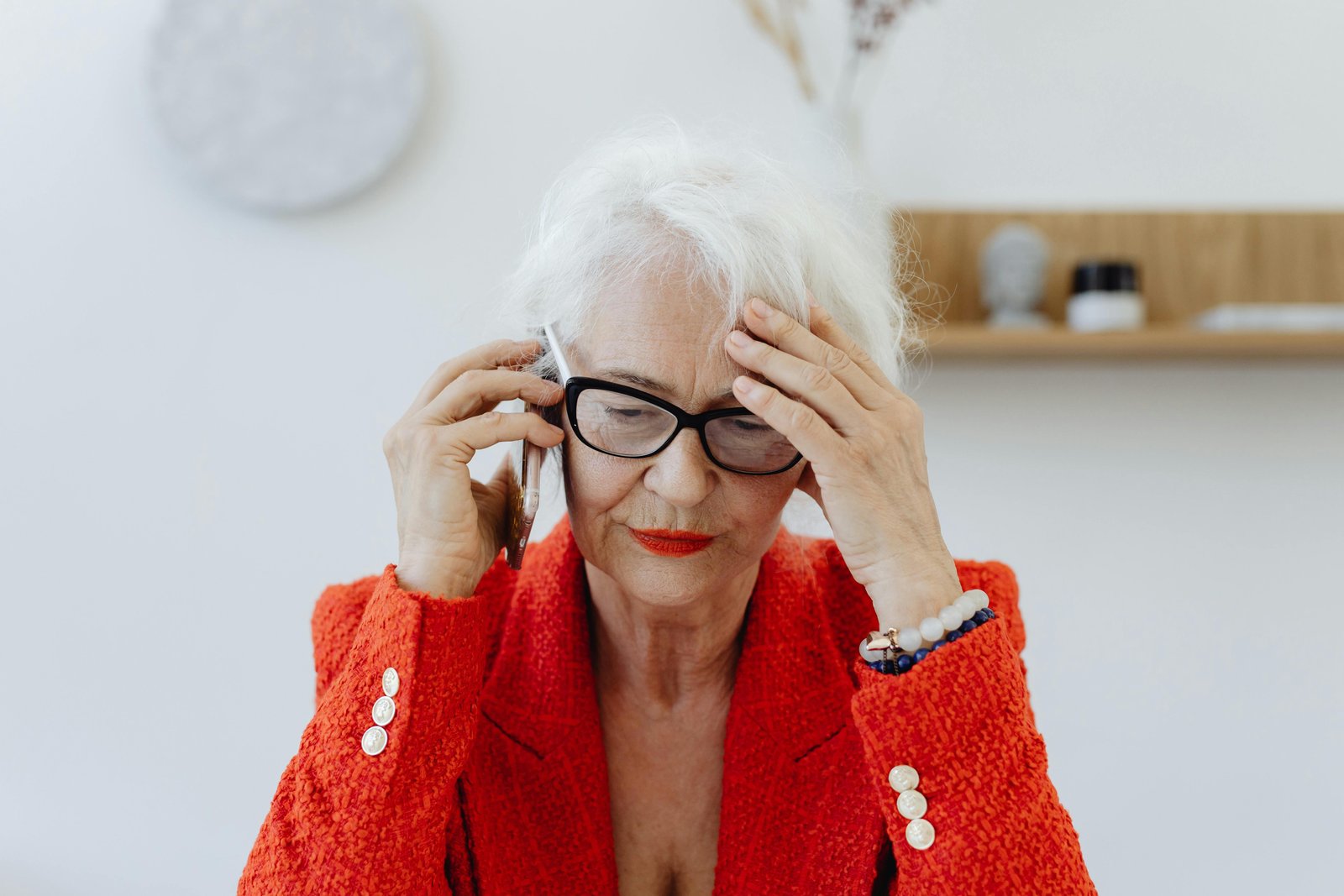 Older woman in a red blazer holding a phone and touching her temple, looking stressed while making a difficult decision