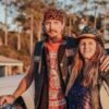 Middle-aged couple standing together outdoors in biker attire, smiling confidently beside a motorcycle during golden hour