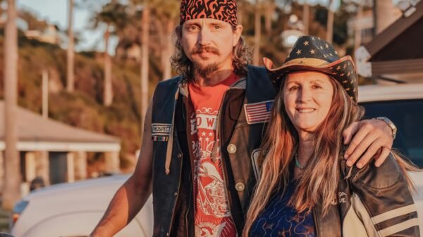Middle-aged couple standing together outdoors in biker attire, smiling confidently beside a motorcycle during golden hour