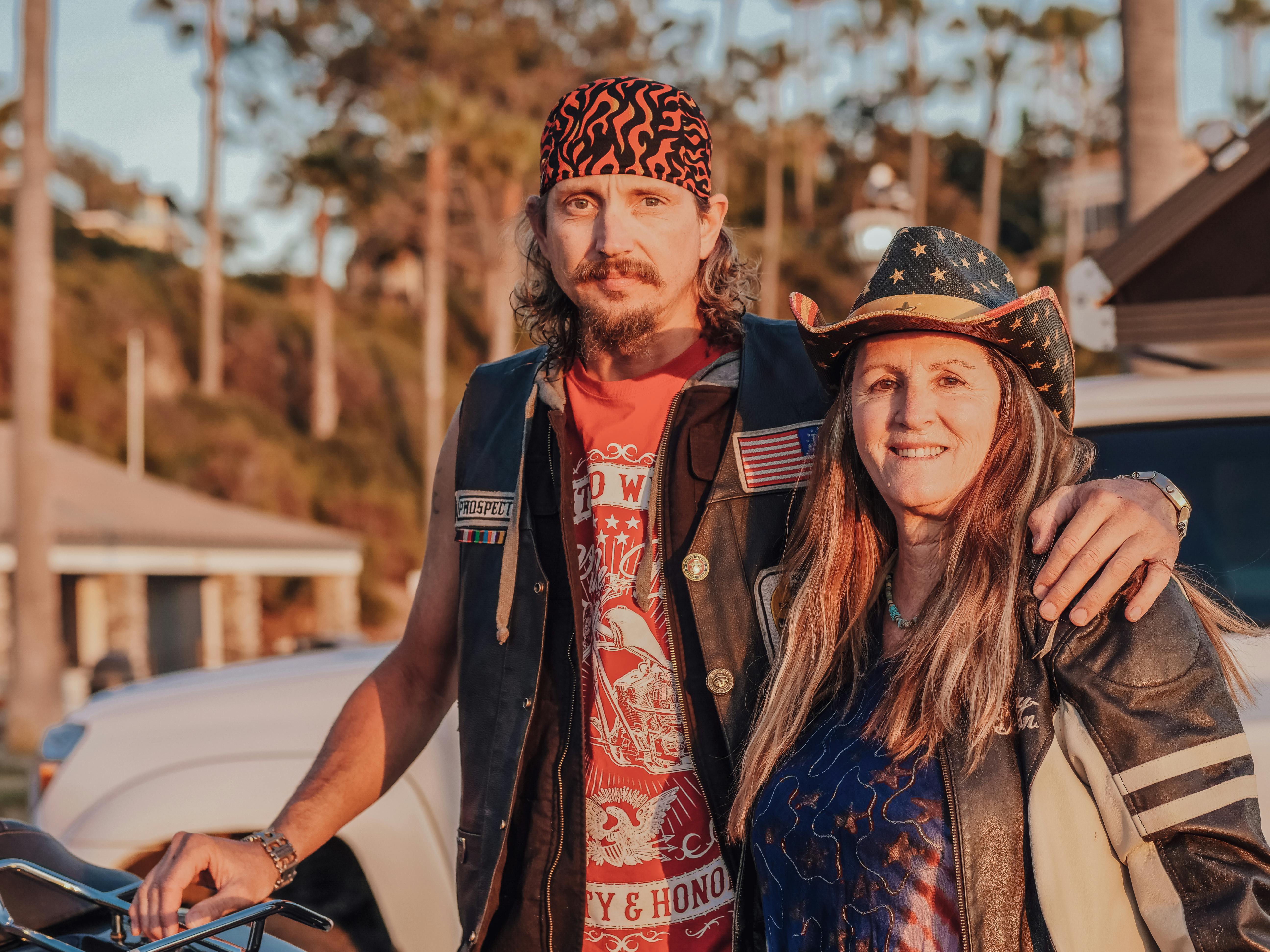 Middle-aged couple standing together outdoors in biker attire, smiling confidently beside a motorcycle during golden hour
