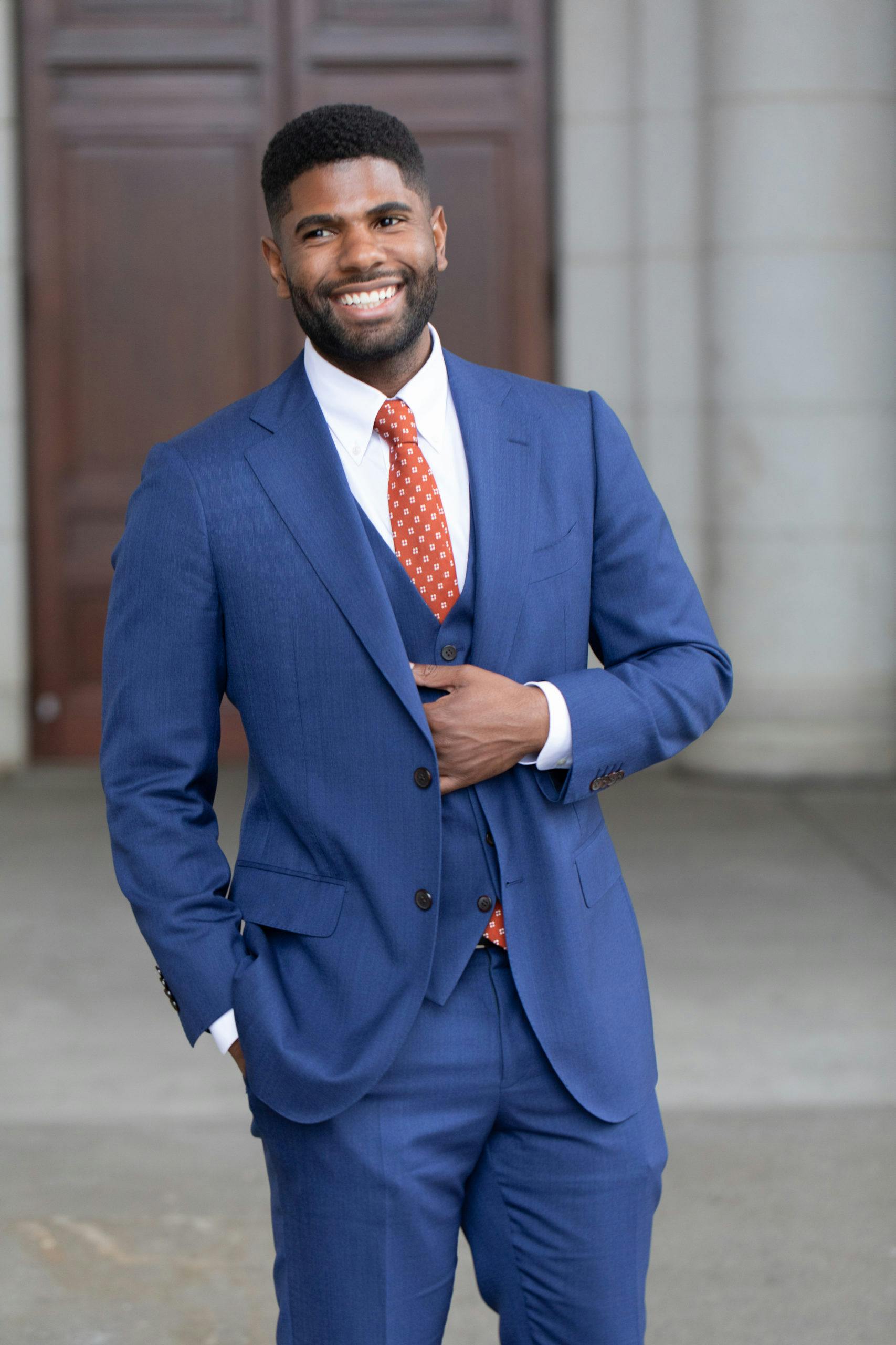 Smiling man wearing a blue suit and red tie, standing confidently outdoors in a professional setting