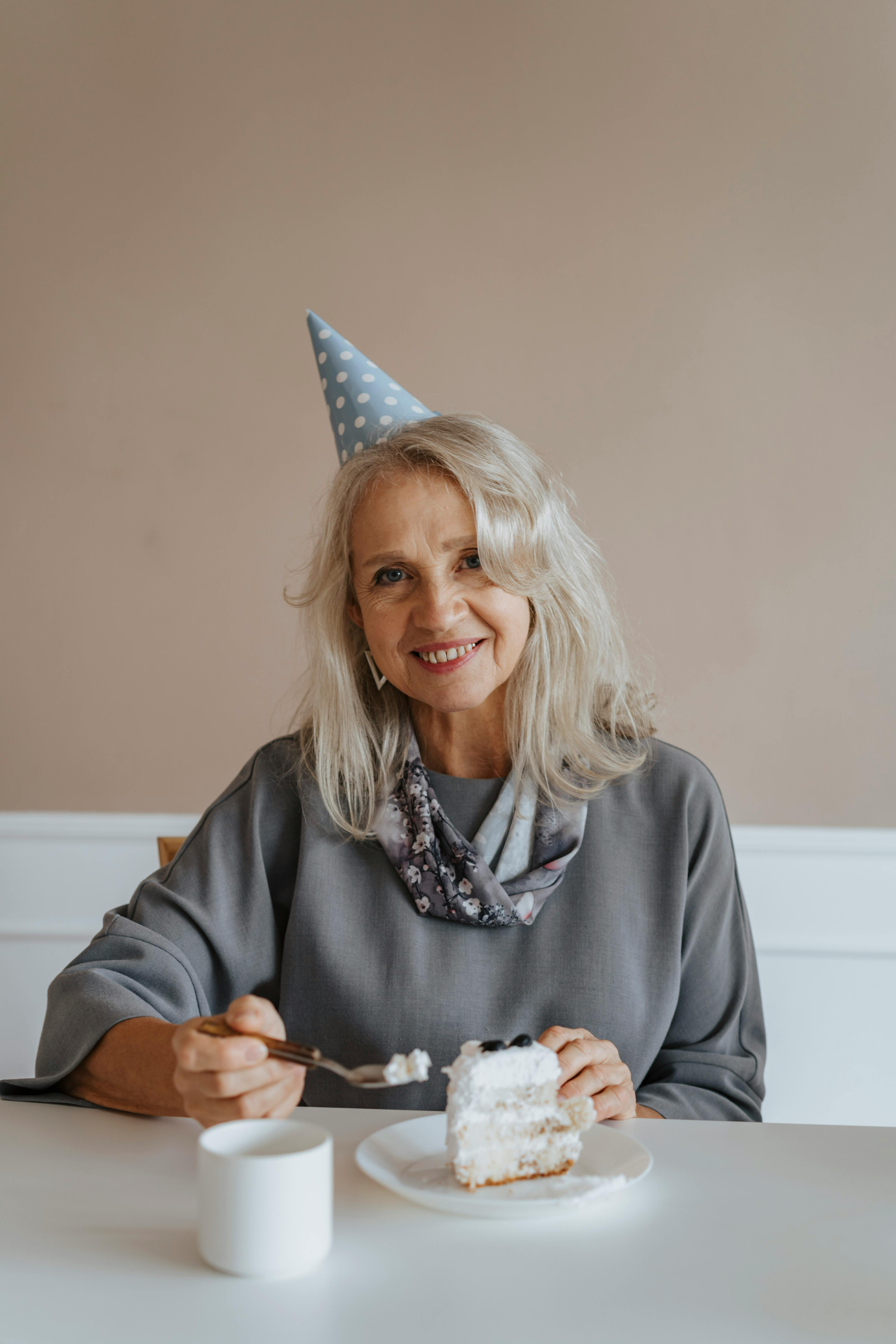 Smiling middle-aged woman wearing a party hat, enjoying a slice of cake at a table