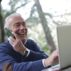 Smiling older man talking on the phone while using a laptop outdoors