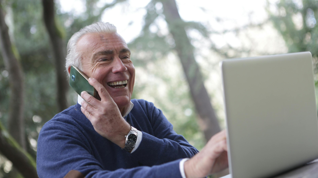 Smiling older man talking on the phone while using a laptop outdoors