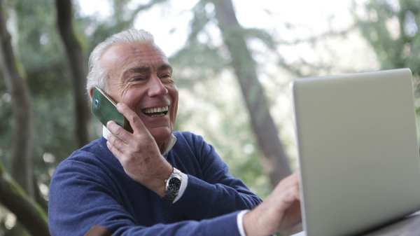 Smiling older man talking on the phone while using a laptop outdoors