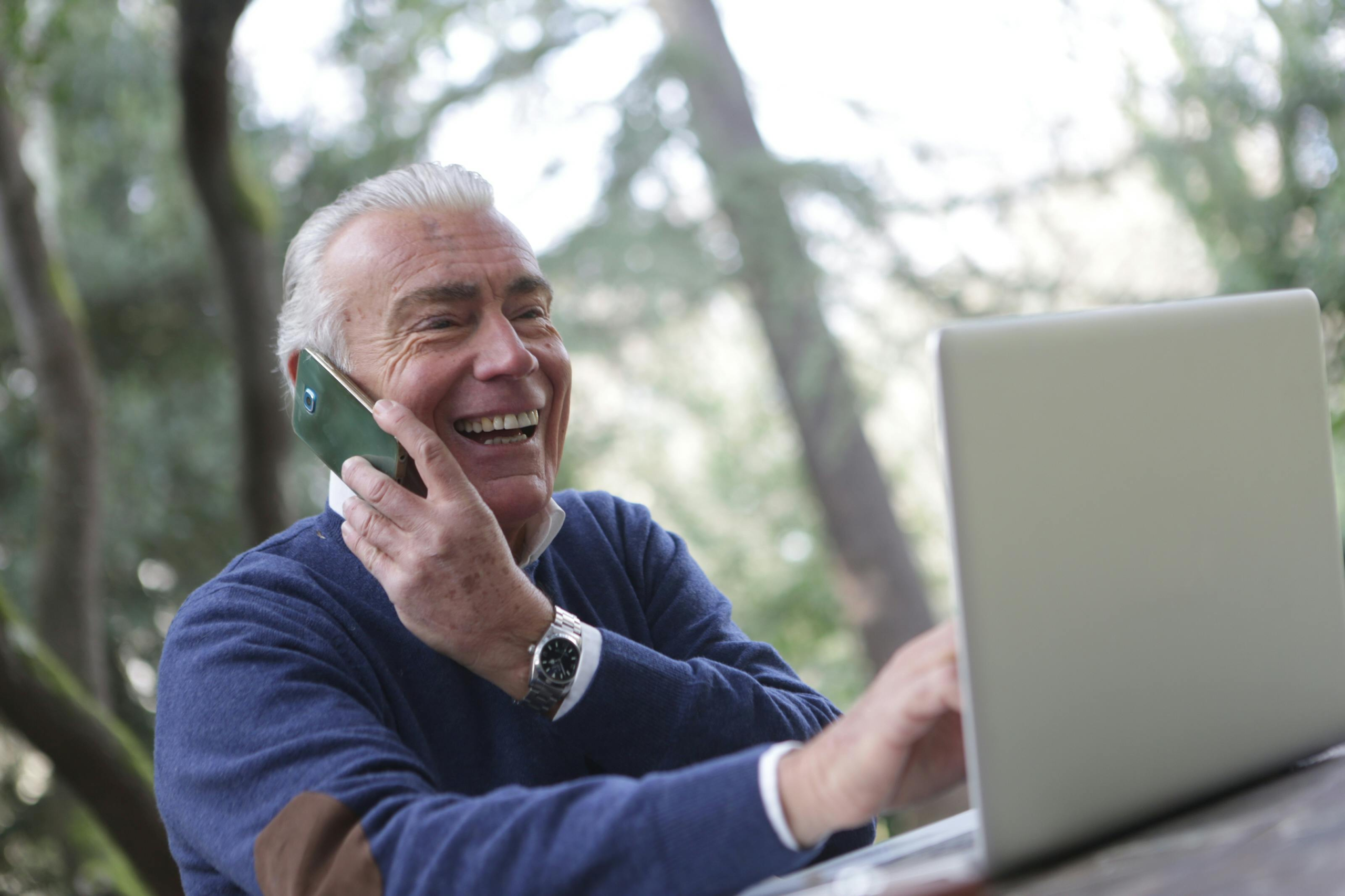 Smiling older man talking on the phone while using a laptop outdoors