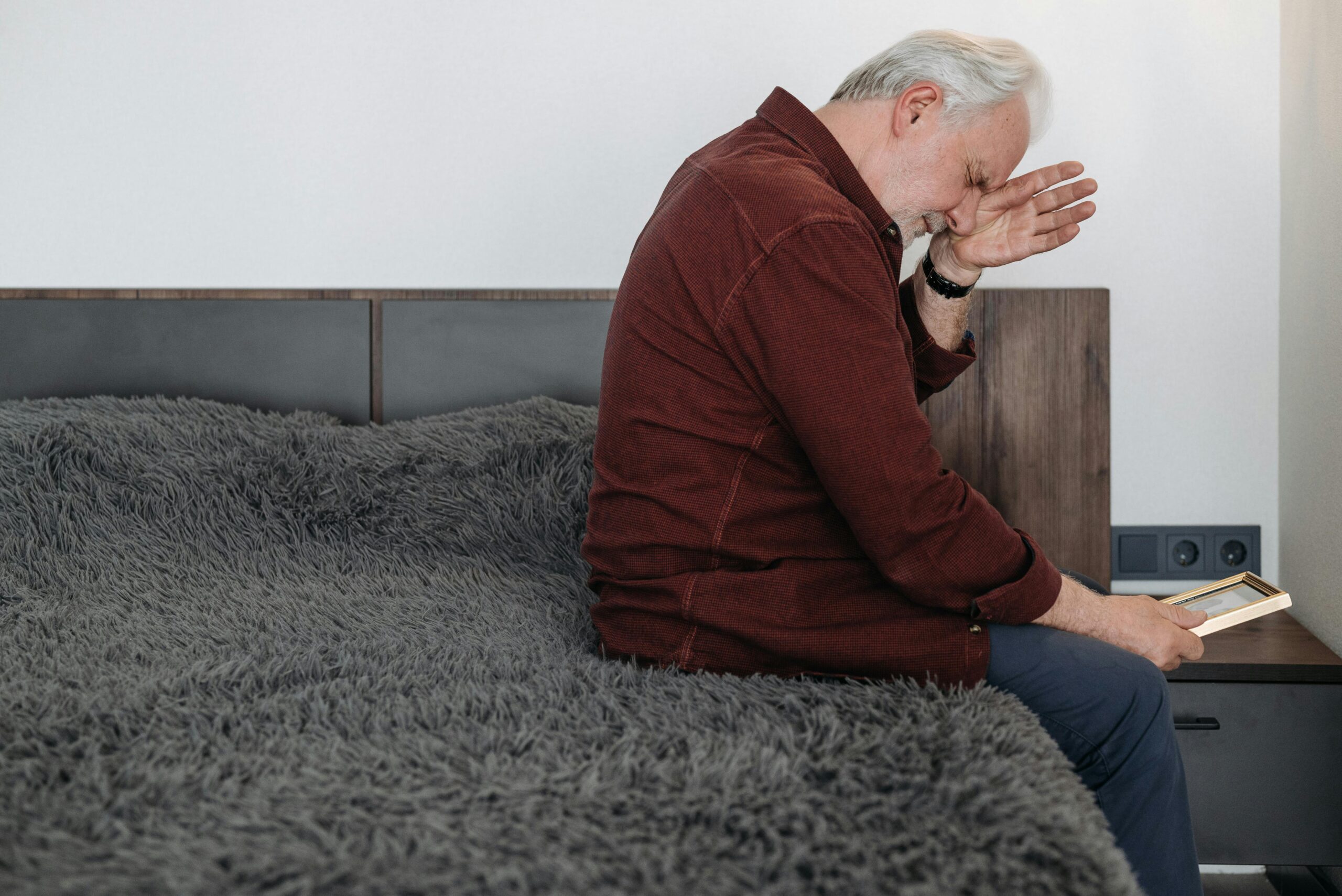 Older man sitting thoughtfully on a bed in a rustic room, reflecting quietly in natural light
