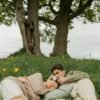 Couple lying together on a blanket in a grassy field beneath trees, sharing a quiet and intimate moment surrounded by nature