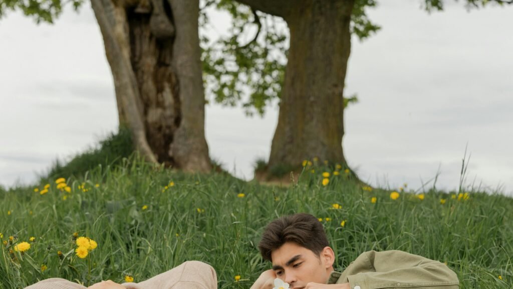 Couple lying together on a blanket in a grassy field beneath trees, sharing a quiet and intimate moment surrounded by nature