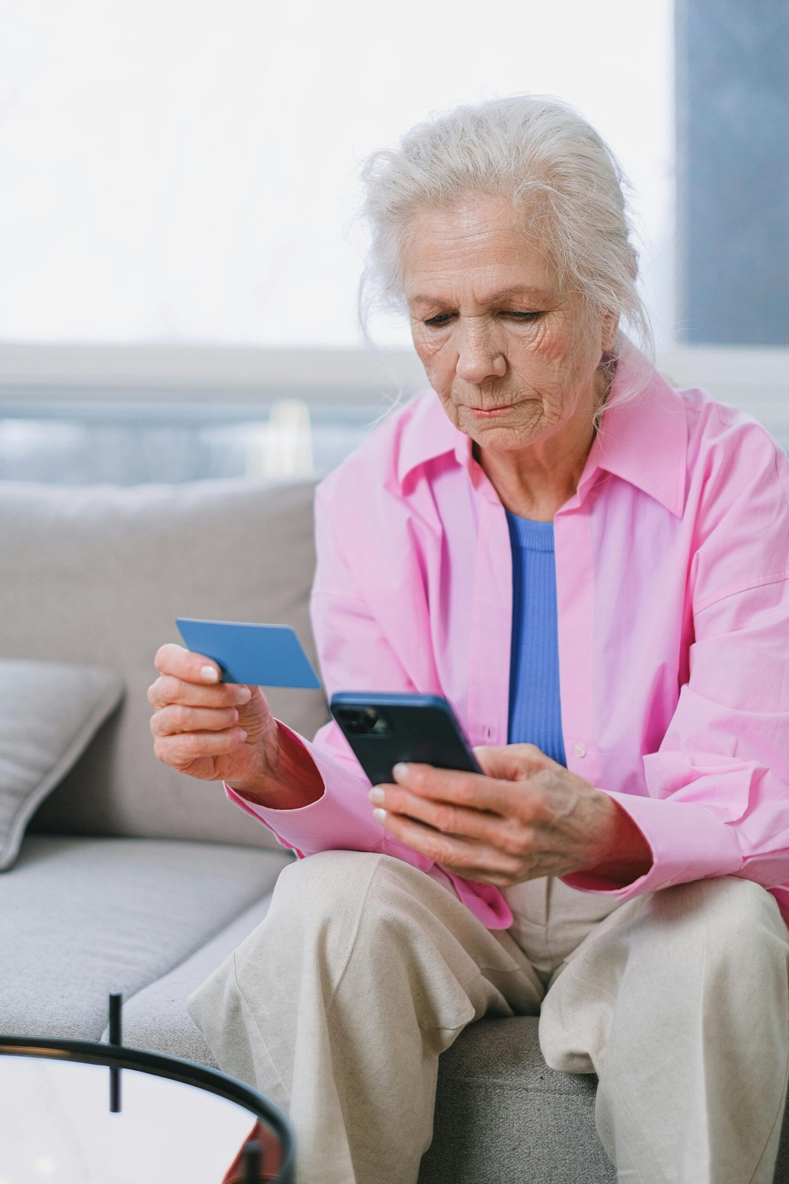 Older woman sitting on a couch holding a credit card and smartphone, focused on managing finances or making an online payment