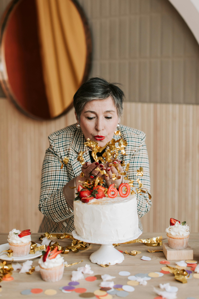 Woman in her 60s blowing gold confetti over a birthday cake, symbolizing reflection and new beginnings
