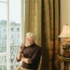 Middle-aged man sitting by a window in a classic room, thoughtfully looking outside while holding a cigarette
