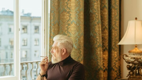 Middle-aged man sitting by a window in a classic room, thoughtfully looking outside while holding a cigarette