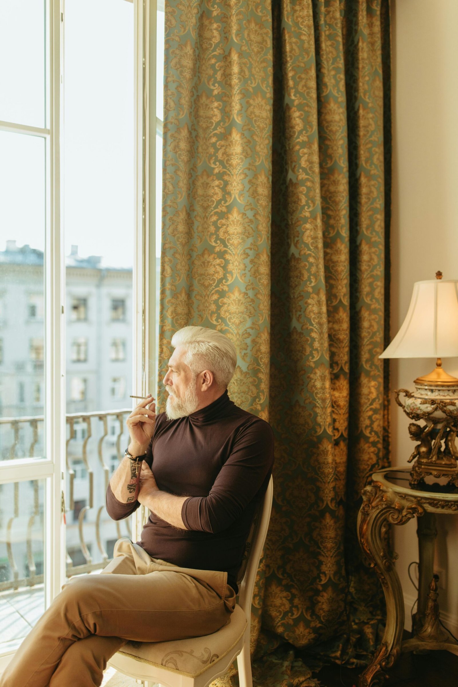 Middle-aged man sitting by a window in a classic room, thoughtfully looking outside while holding a cigarette