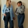 Two midlife women standing together indoors, smiling and talking, dressed in casual modern outfits against a neutral wall