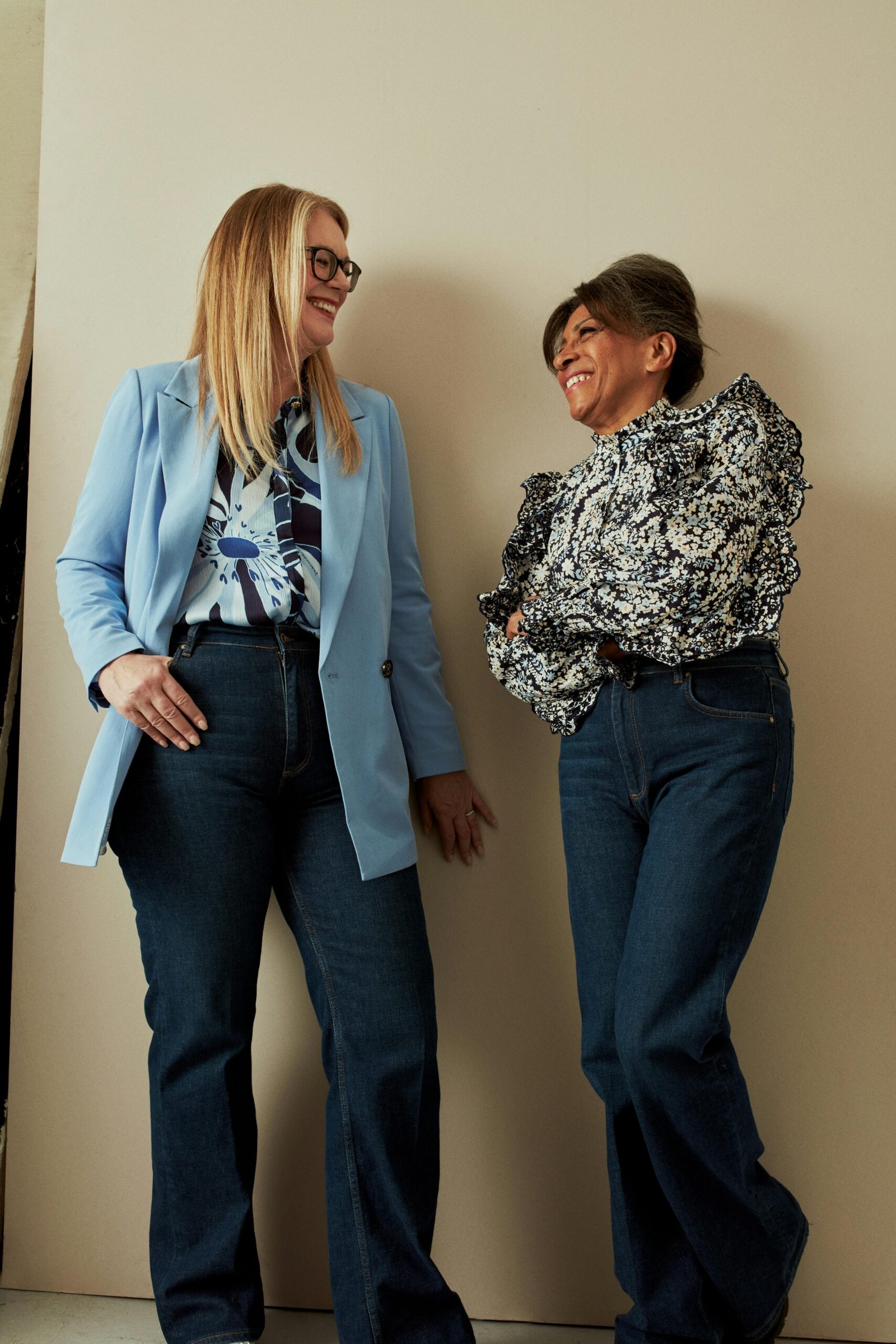 Two midlife women standing together indoors, smiling and talking, dressed in casual modern outfits against a neutral wall