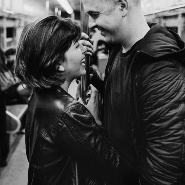 Black and white photo of a couple smiling at each other on a subway train