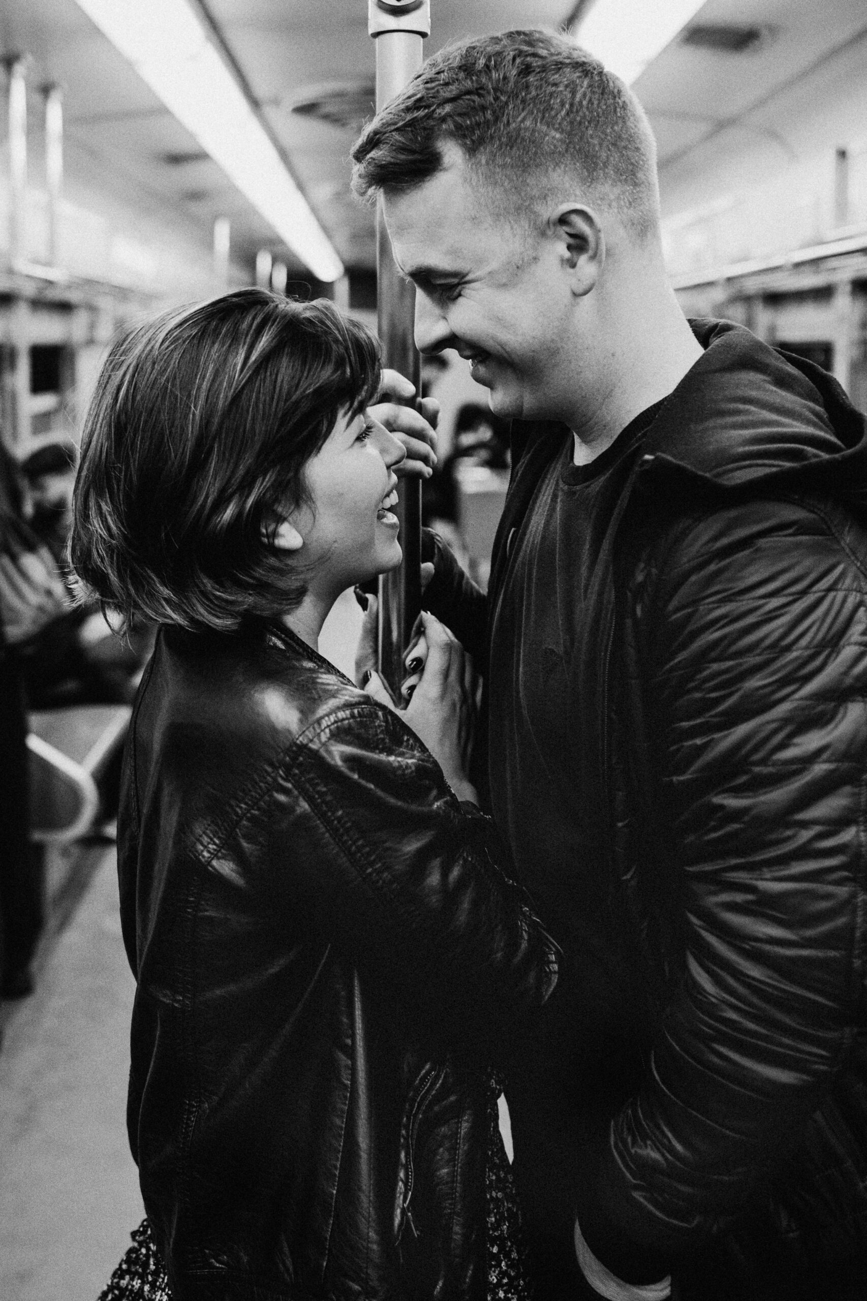 Black and white photo of a couple smiling at each other on a subway train