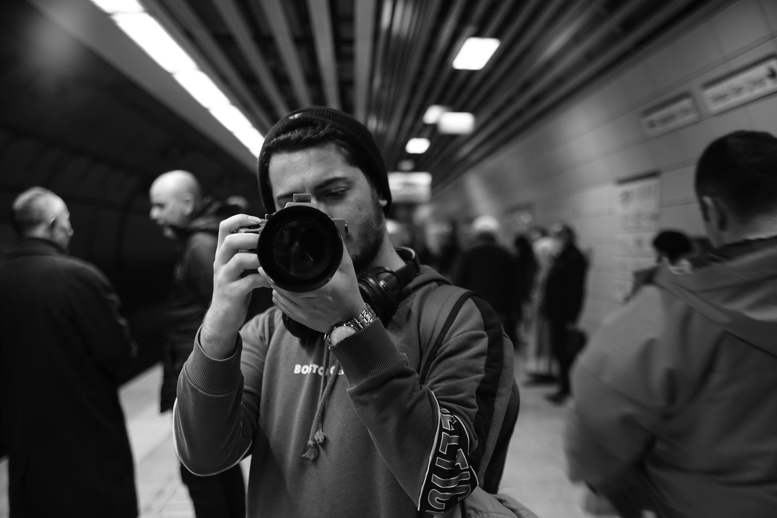 Person taking a photograph in a busy subway station, surrounded by commuters, captured in black and white